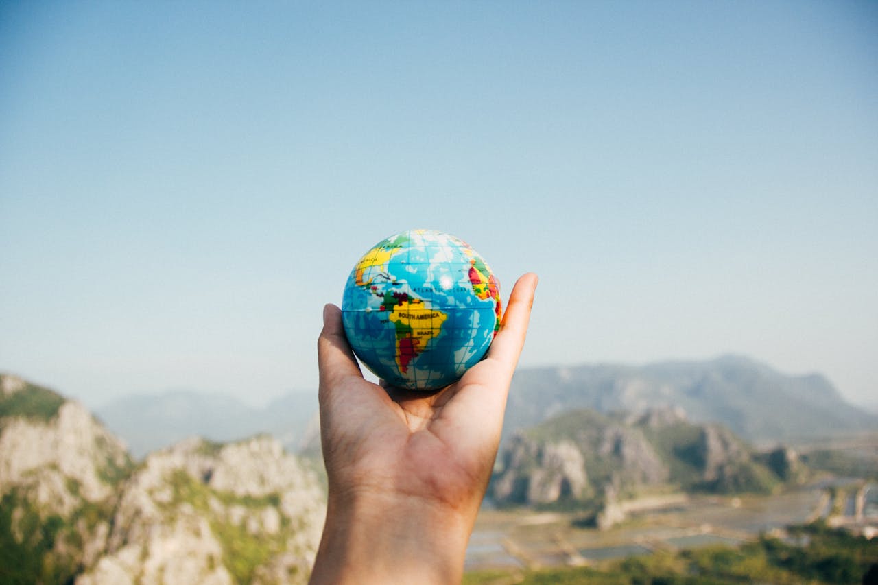 heros-img Hand holding a globe against a mountain background symbolizing travel and exploration.